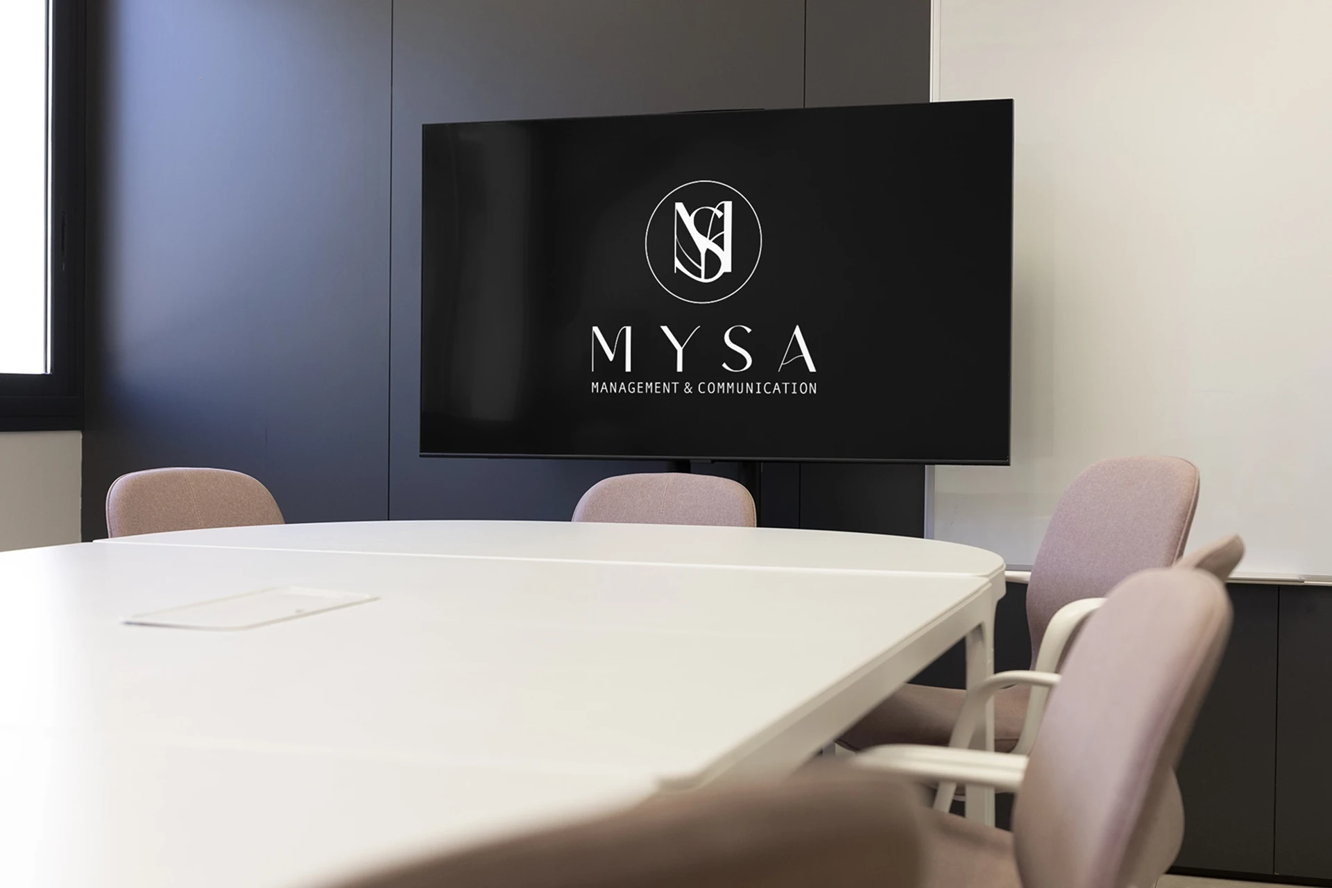 The MYSA logo on a wall-mounted screen in a meeting room with blush chairs and a white table